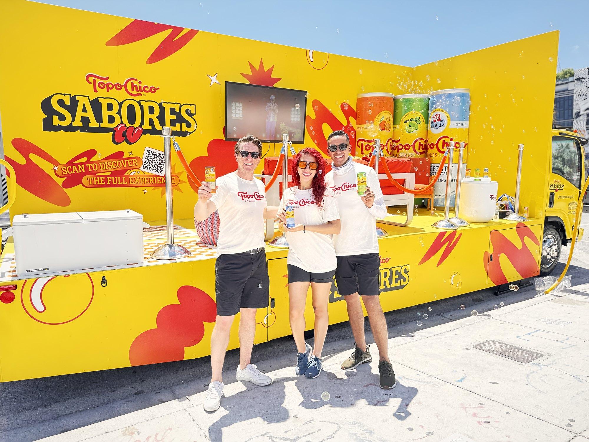 Three ambassadors raise drinks in front of a bright yellow mobile stage truck with lounge seating
