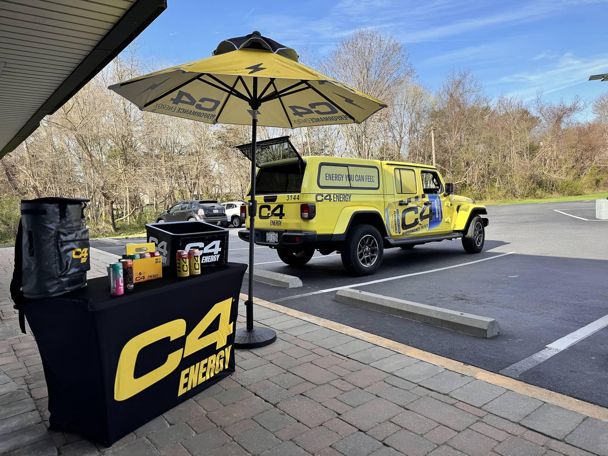 Branded sampling table and umbrella beside a wrapped tour vehicle in a sunny parking lot