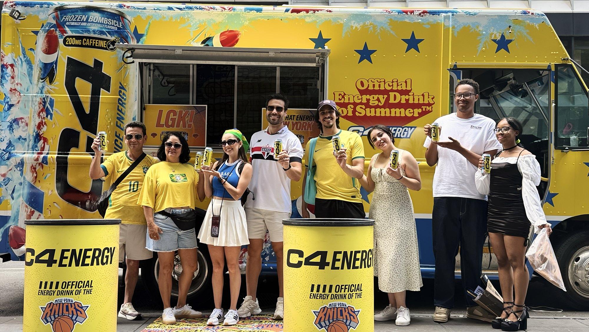 Smiling fans in soccer jerseys hold energy drink cans in front of a yellow summer sampling truck