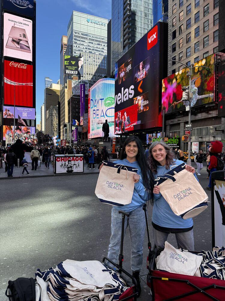 Two street team members in matching blue hoodies hold up branded tote bags in Times Square