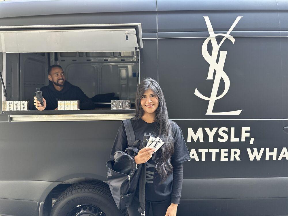A brand ambassador smiles beside a black fragrance tour van while a teammate hands out samples from the window