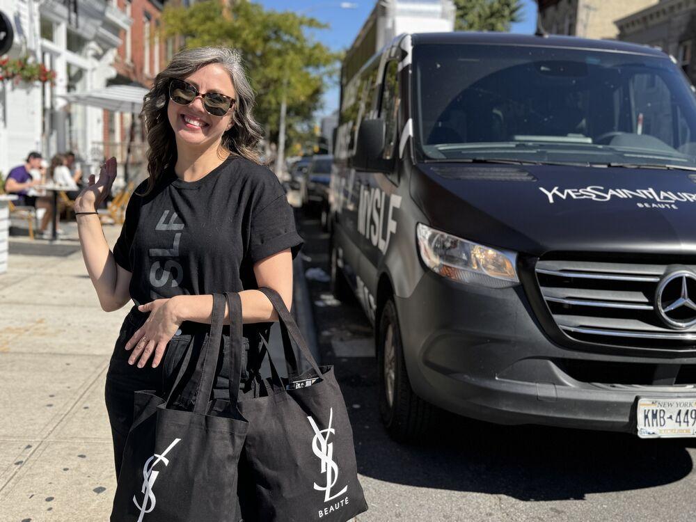 An ambassador with branded tote bags waves beside a black tour van on a neighborhood street