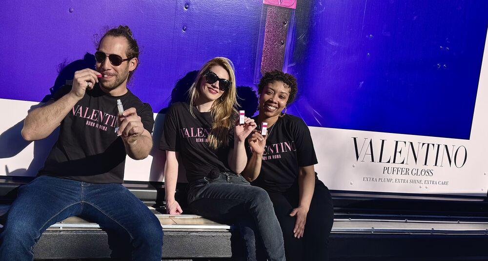 Three ambassadors in black tees try lip gloss samples on the step of a purple wrapped tour truck