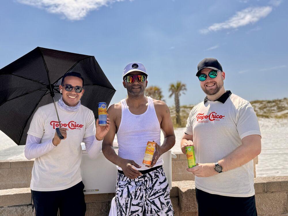 Three ambassadors hold sparkling water cans by the dunes on a sunny beach day